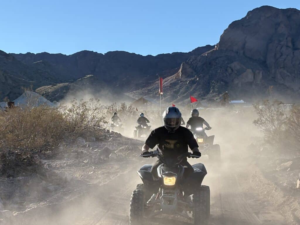 ATV riders explore a dusty Nevada desert trail near Las Vegas with rocky hills and mountains under a clear blue sky.