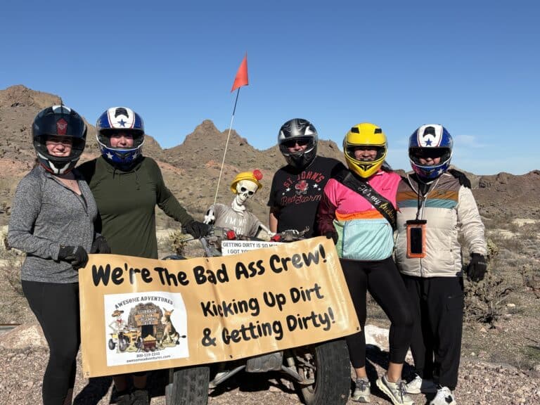 ATV riders in helmets pose in Nevada desert near Las Vegas with a fun banner and skeleton, ready for thrilling RZR off-road adventures.