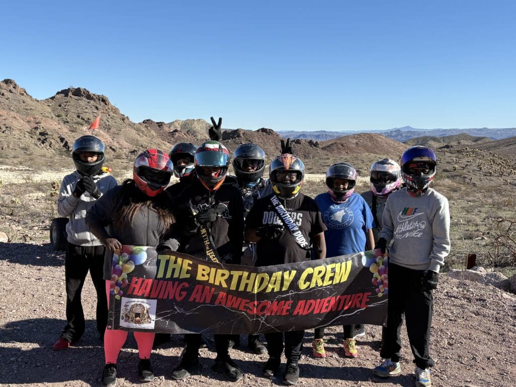 Adventurers in helmets pose in Nevada’s rocky desert with “The Birthday Crew” banner after an epic Las Vegas ATV tour.