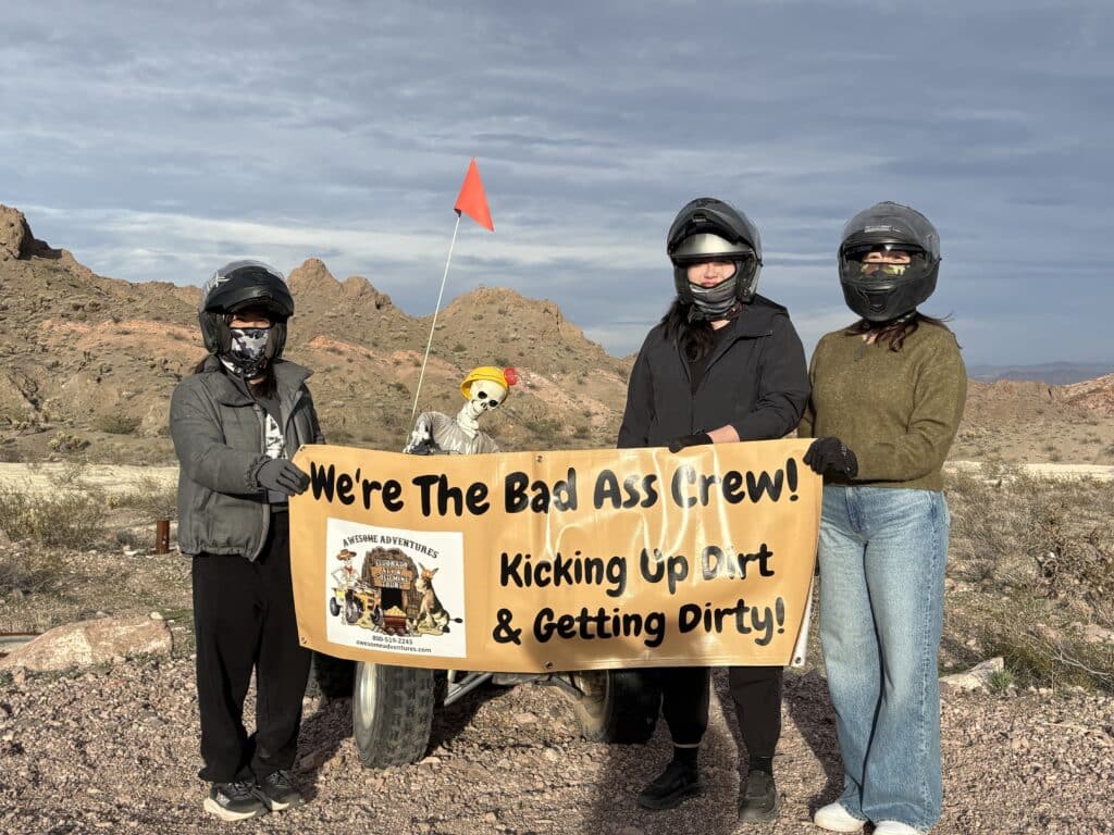 Three with helmets and skeleton on ATV hold “Bad Ass Crew” banner in Nevada desert, near Colorado River—ATV tour adventure!.