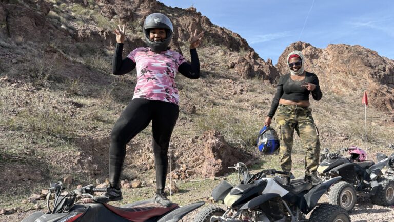 Two women on ATVs pose with peace signs during a Nevada ATV tour near rocky hills, perfect for Las Vegas off-road adventures.