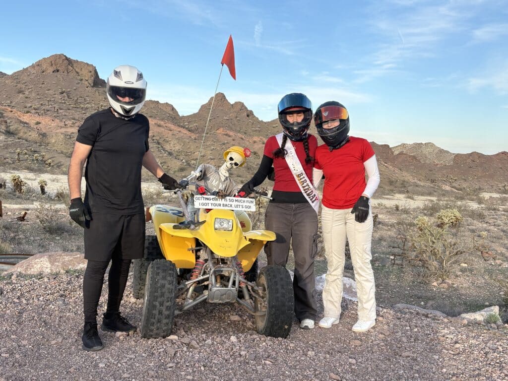 Three riders in helmets by a yellow ATV and skeleton in Nevada desert, perfect for Las Vegas ATV tours and Ghost Town adventures.