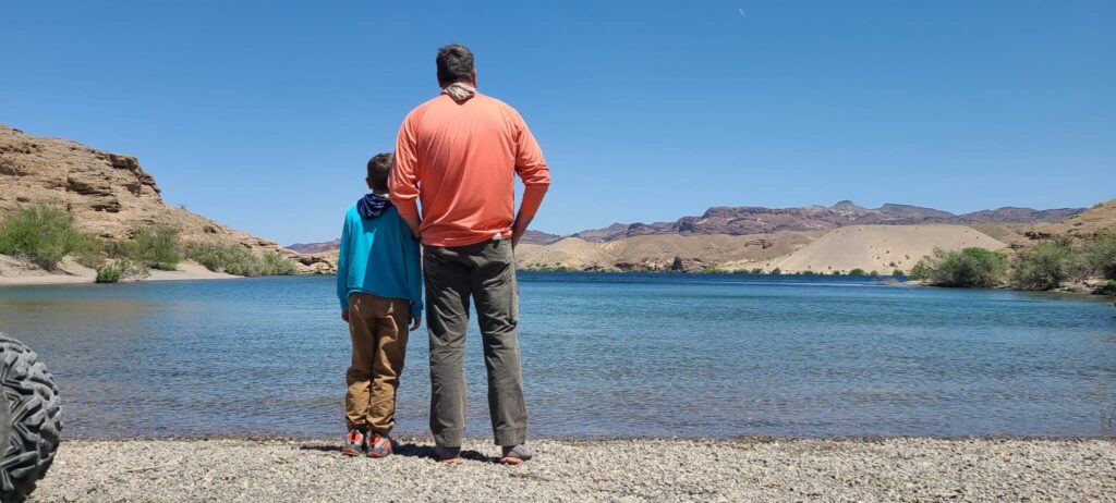 Father and child by Colorado River near Las Vegas, Nevada, after an exciting Ghost Town ATV tour with desert hills in the background.