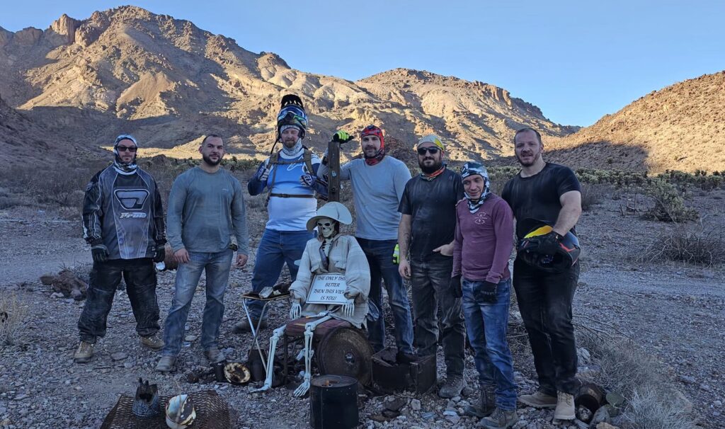 Seven men in off-road gear join a skeleton on a Las Vegas Ghost Town ATV tour, rocky Nevada desert and mountains behind.