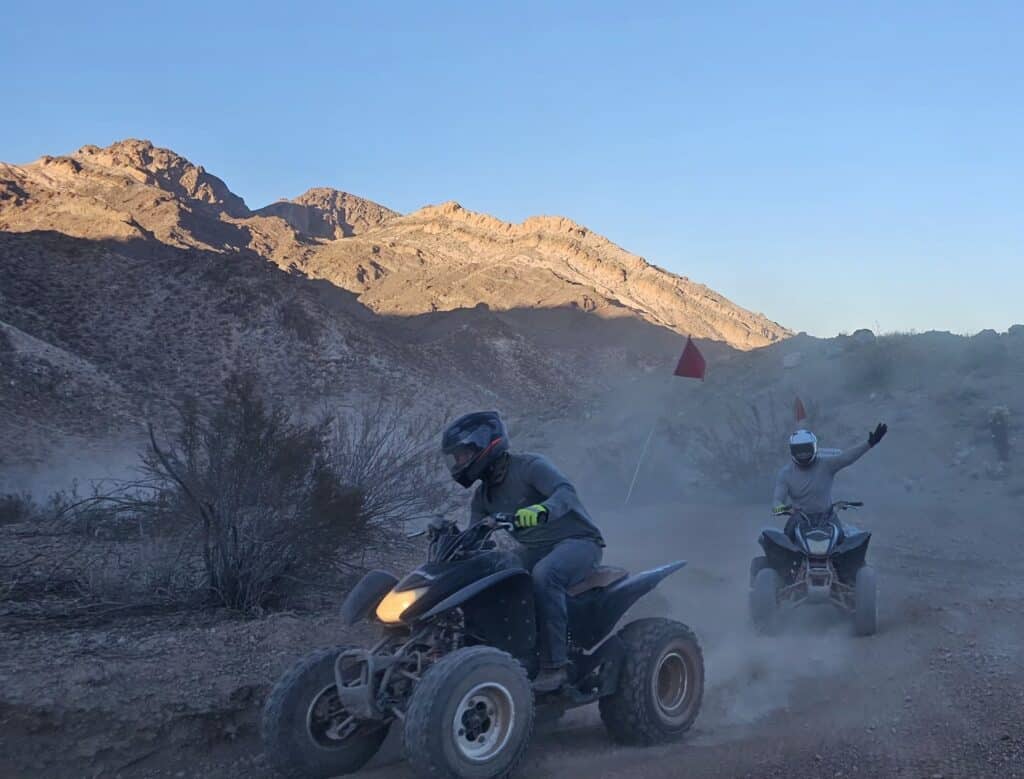Adventurers ride ATVs on a dusty Nevada trail near Las Vegas, exploring Eldorado Canyon and waving under sunny desert skies.