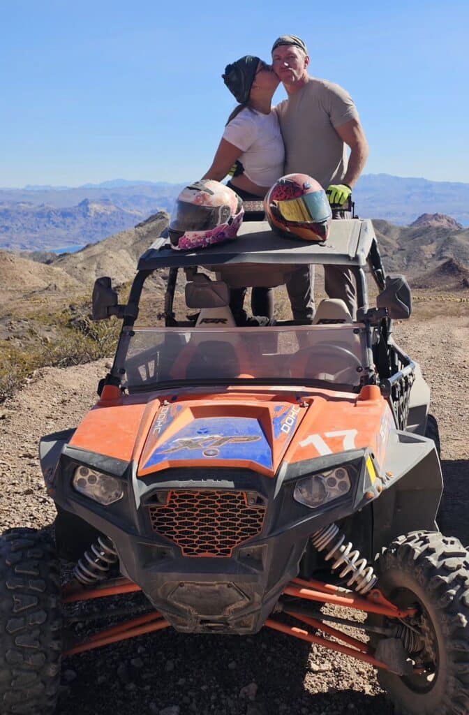 Couple kisses on orange ATV in Las Vegas, Nevada, with helmets, mountains, Colorado River—perfect for RZR off-road tours.