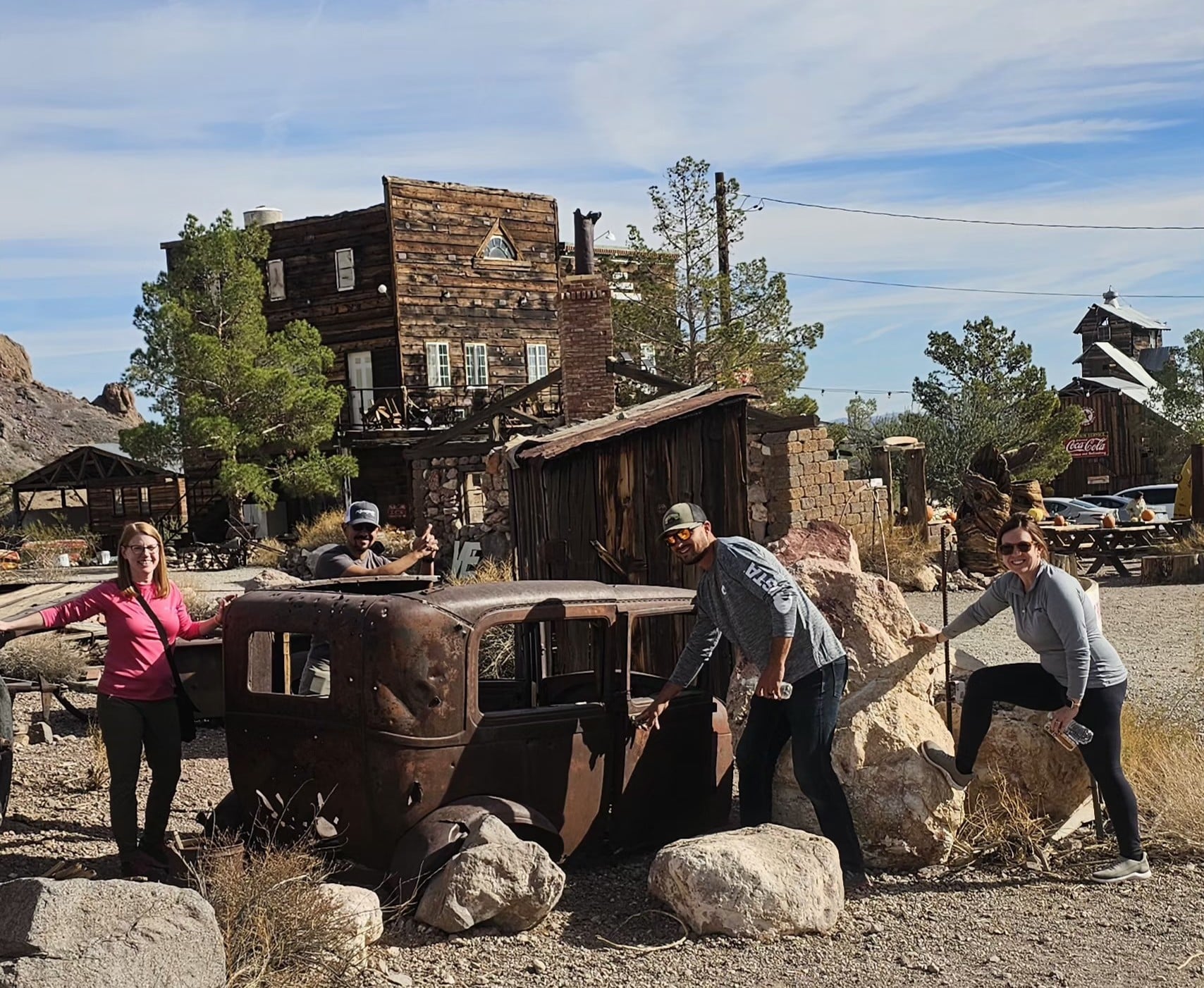 Group posing by a rusty car at a Nevada ghost town on a Las Vegas ATV tour, wooden buildings and clear sky in the desert.