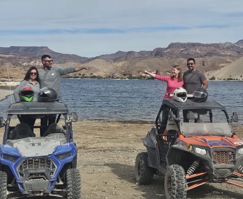 Las Vegas RZR ATV tour group smiles by off-road vehicles on a Nevada desert lake shore near the Colorado River.