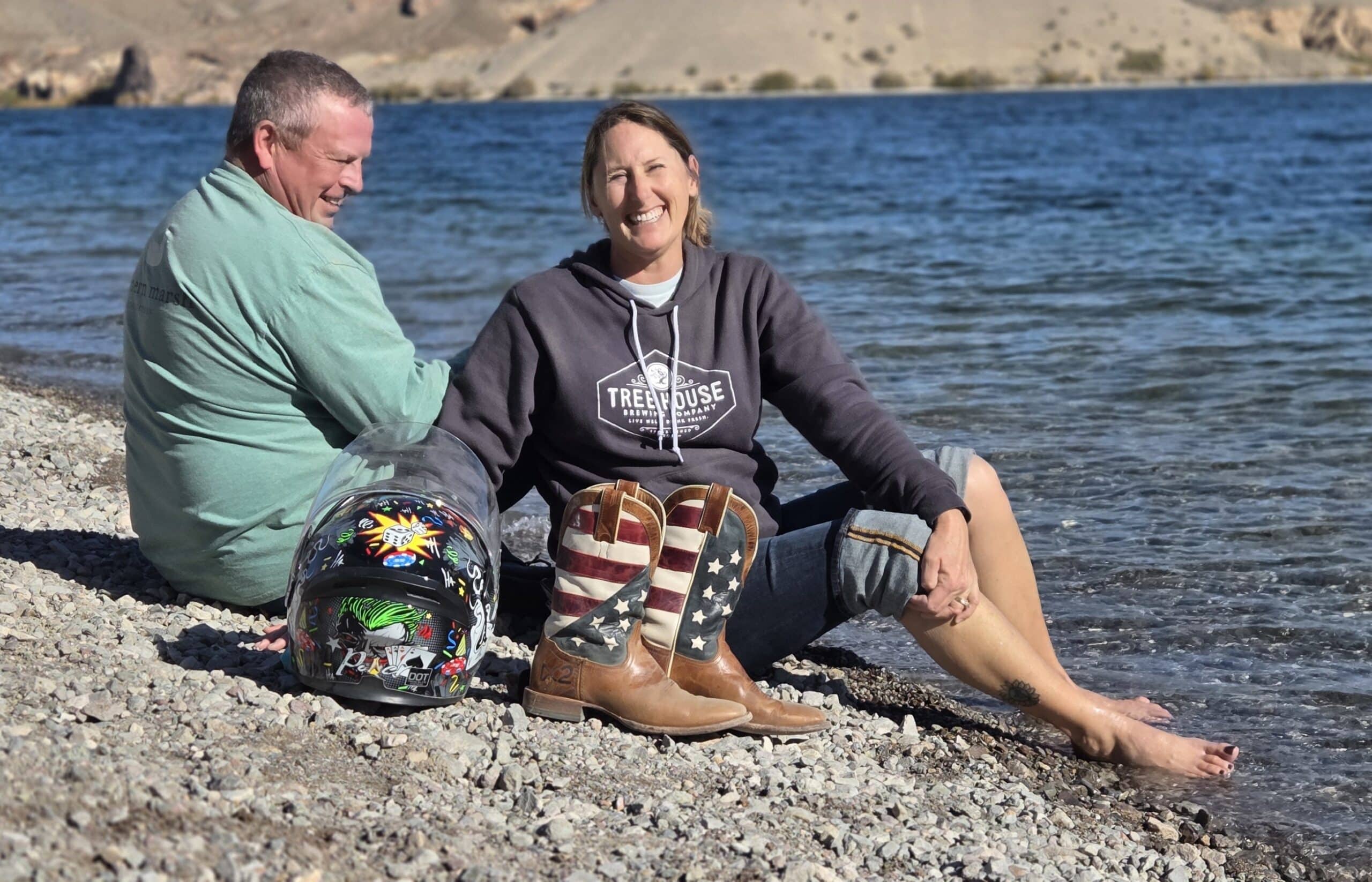 Smiling couple relaxes lakeside near Las Vegas after a Nevada RZR tour; helmet, flag boots, desert hills & Colorado River shown.