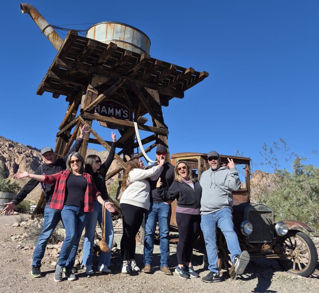 Group poses by an old water tower and vintage car, enjoying Las Vegas ATV tours near Nevada’s rocky hills under a clear sky.
