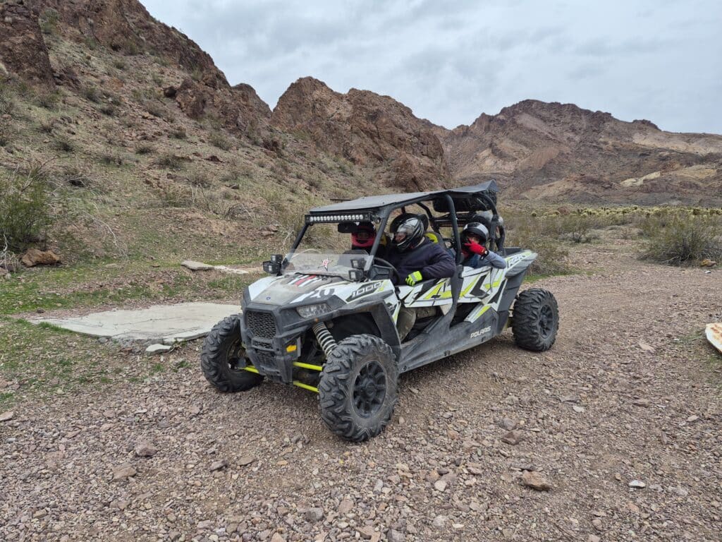 Two people in helmets ride a gray and yellow RZR during an ATV tour near Las Vegas, Nevada desert mountains under cloudy skies.
