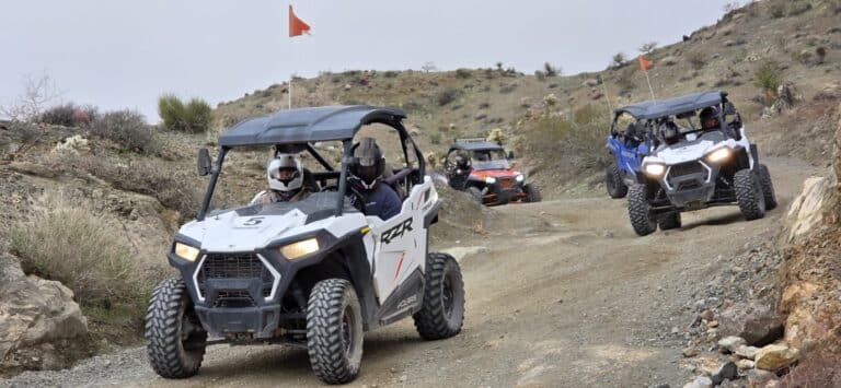 ATV tour near Las Vegas: three off-road vehicles with flags ride rocky Nevada desert trails, drivers in helmets, sparse hills.