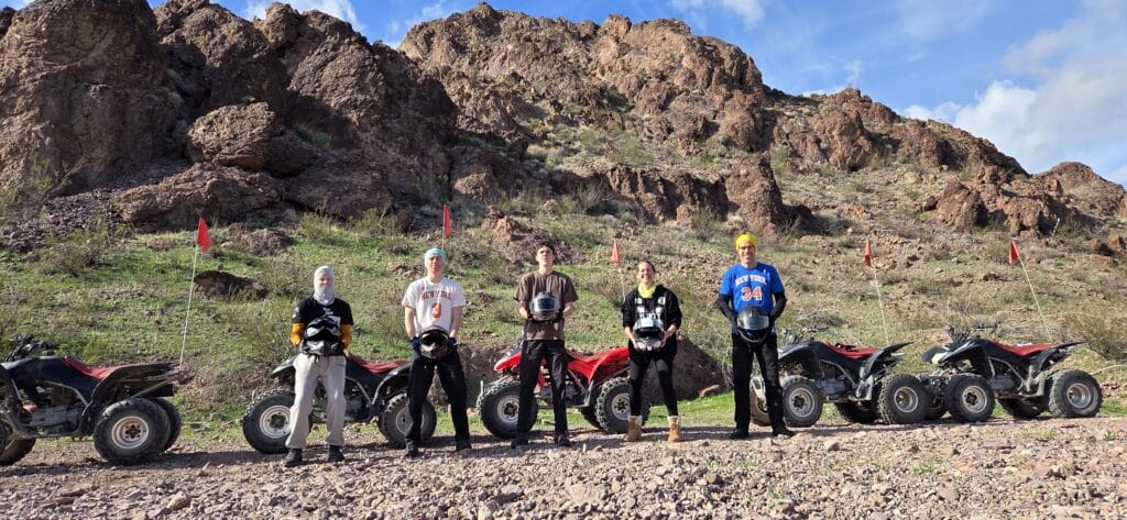 Group in protective gear poses by ATVs with red flags amid Nevada’s rocky terrain—ATV tour near Las Vegas & Colorado River.
