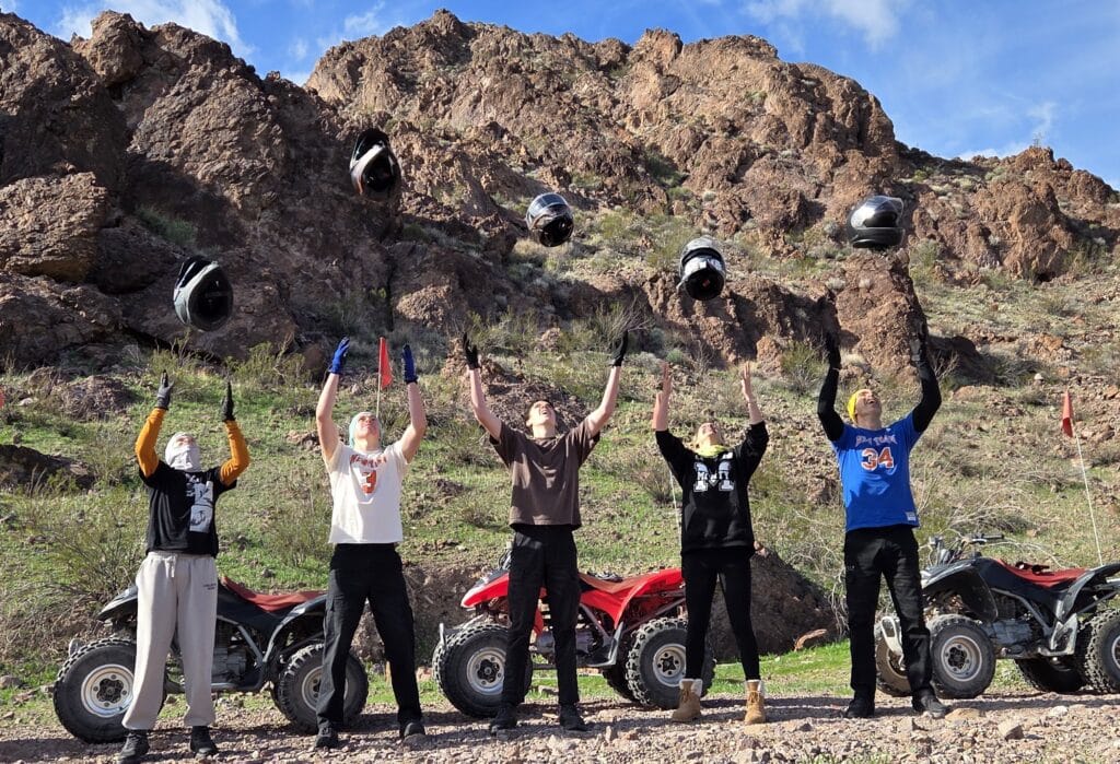Group celebrates an ATV tour near Las Vegas, Nevada, tossing helmets in the air by rocky hills under a clear blue sky.
