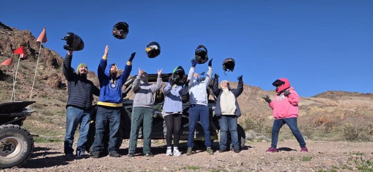 Group celebrates tossing helmets after an ATV tour near Las Vegas, Nevada, with rocky hills and clear skies in the background.