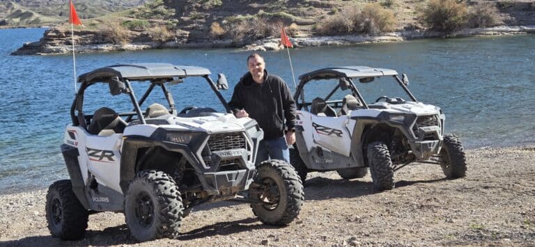 Smiling person between two white off-road vehicles by the Colorado River near Las Vegas, Nevada; both with red flags.