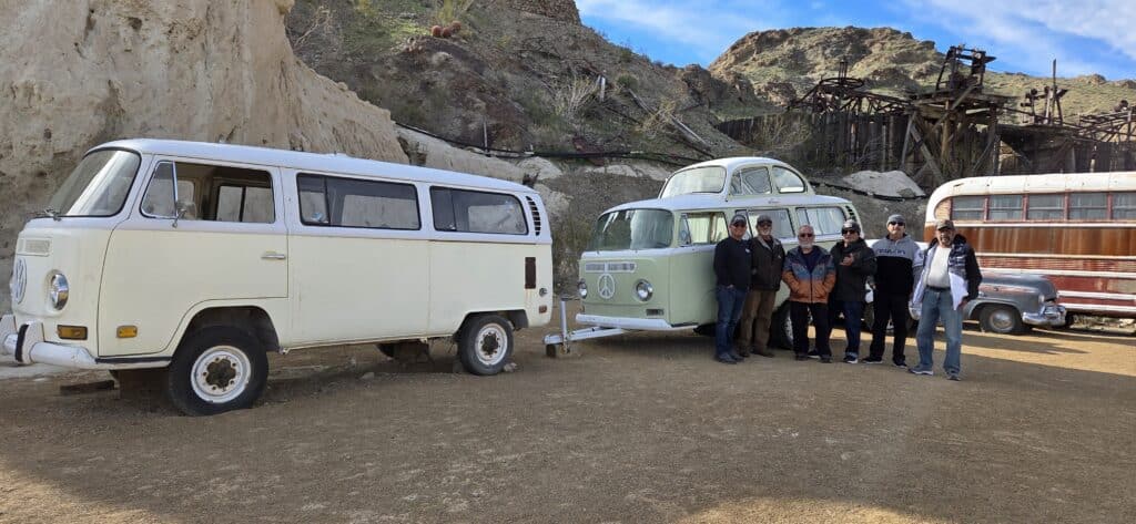 Group by vintage Volkswagen van in Nevada desert near Las Vegas, rocky hills, ghost town sights, and classic trailer in view.