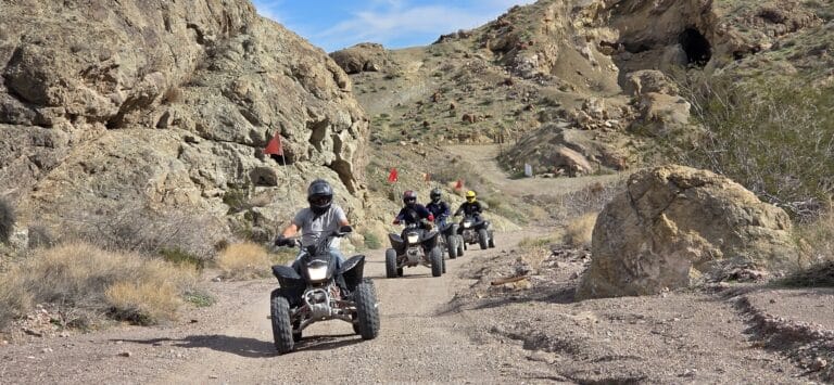 ATV riders explore a rugged Nevada desert trail near Las Vegas, with cliffs and sparse vegetation under a partly cloudy sky.
