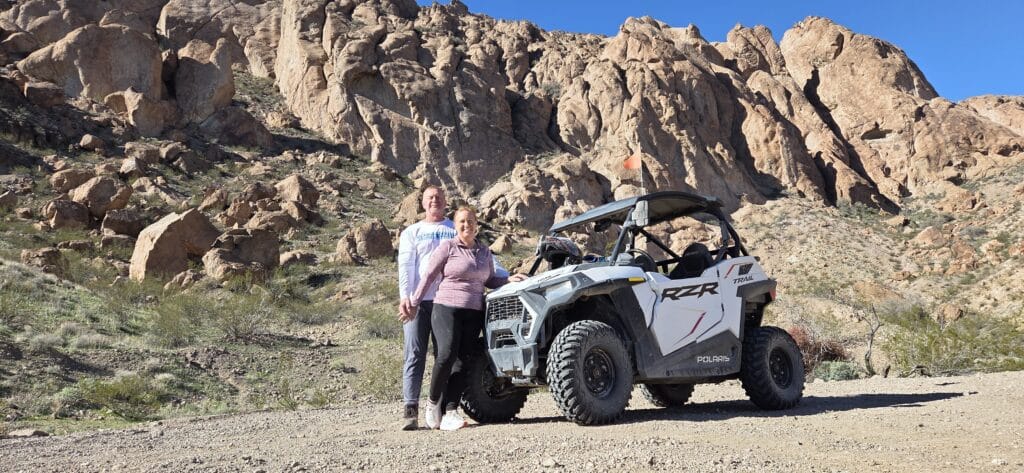 A man and woman stand by a white Polaris RZR during an ATV tour near Las Vegas, Nevada, with rocky hills and blue sky behind.