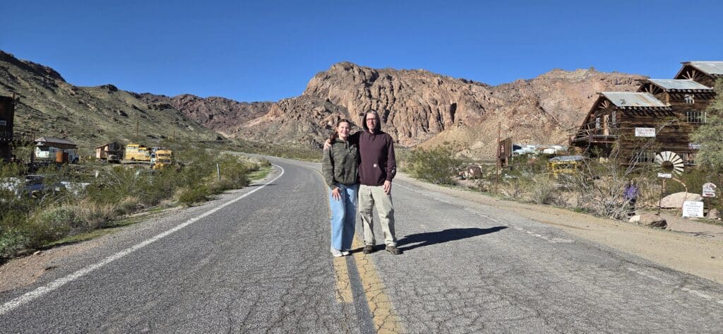 Smiling friends on a Nevada desert road near Las Vegas, with rustic buildings and mountains—perfect for Ghost Town sightseeing.