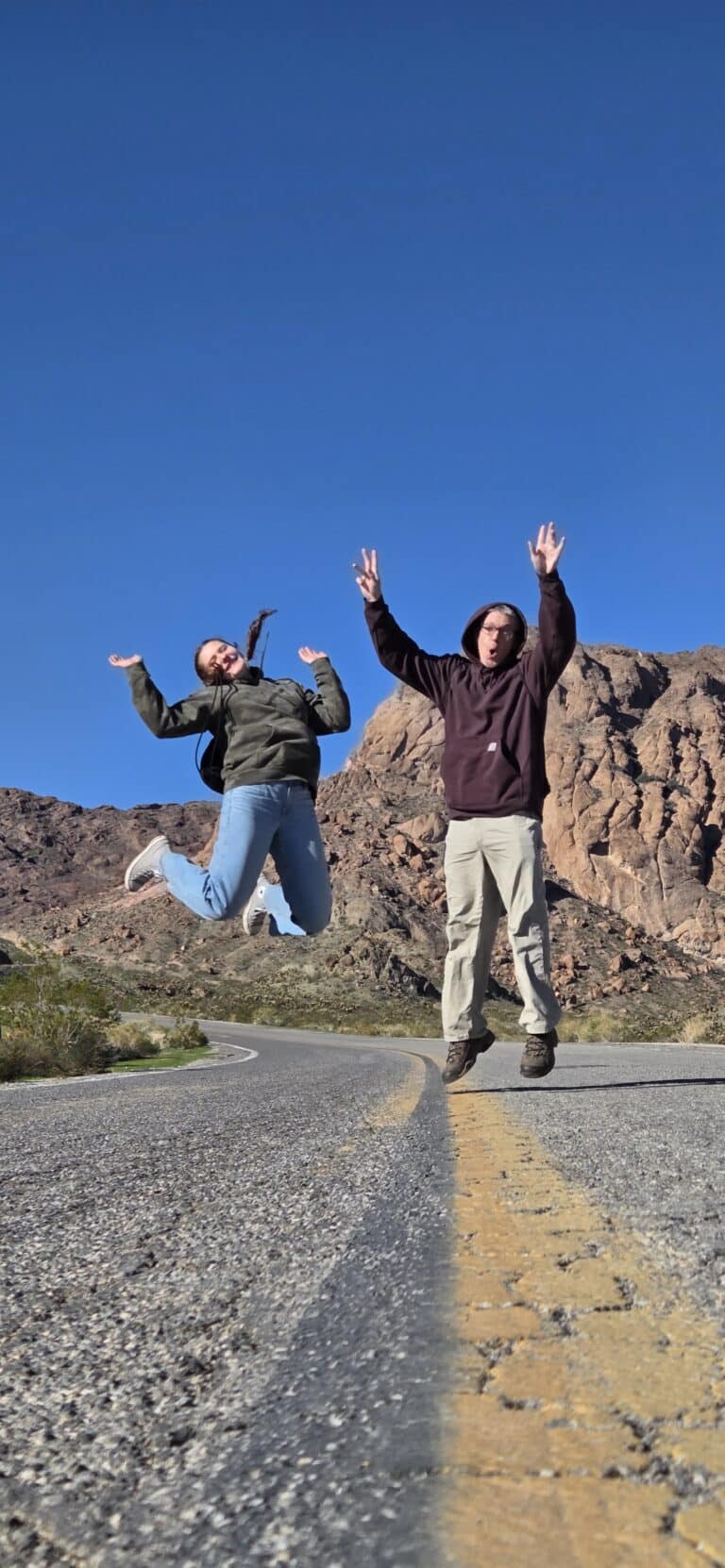 Two people joyfully jump on an empty Nevada road near mountains, perfect for Las Vegas ATV tours or Colorado River adventures.
