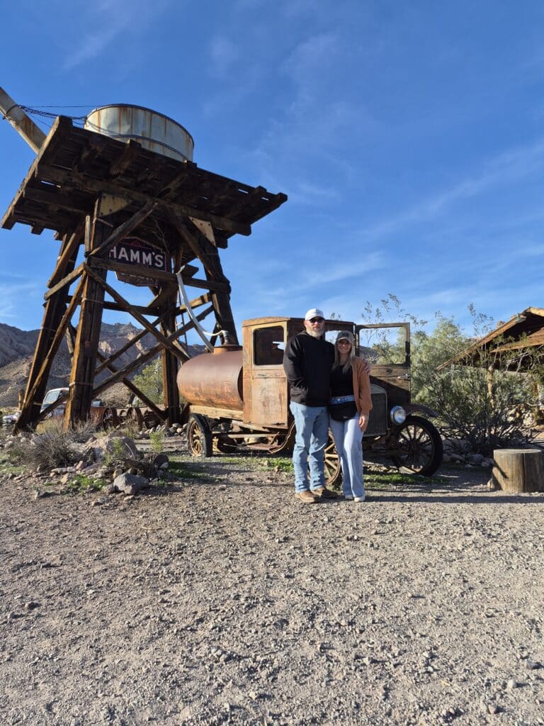 Smiling couple by vintage truck and HAMMS water tower in a Nevada desert ghost town near Las Vegas; perfect for ATV tours.
