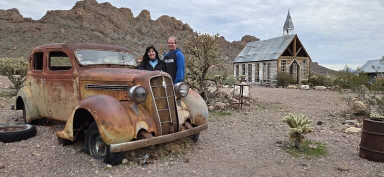 Smiling couple in Nevada ghost town near Las Vegas, standing by vintage car with rocky hills and wooden church, perfect for sightseeing.