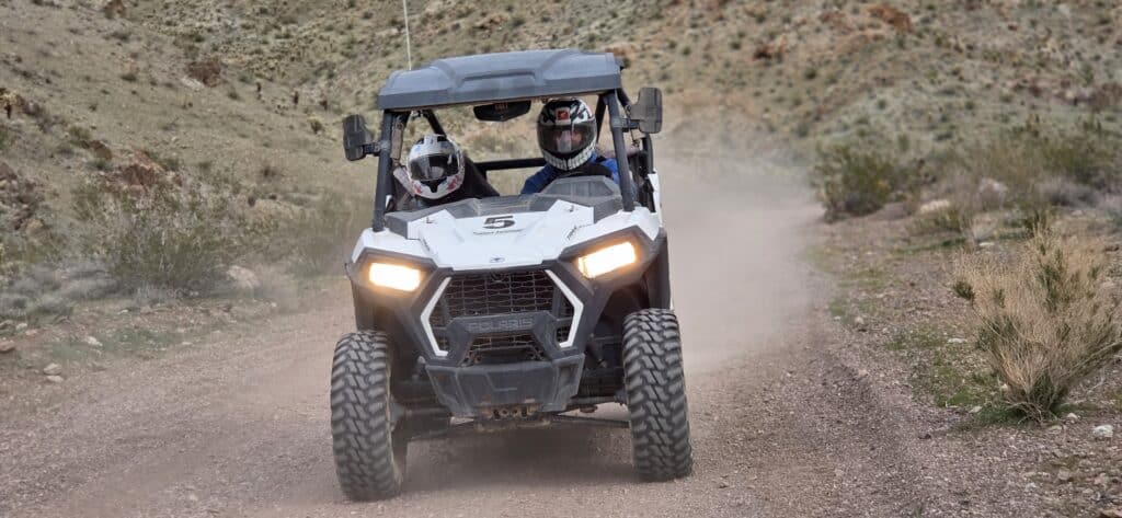 Two helmeted riders on a white RZR ATV tour Nevada’s dusty desert trail near Las Vegas, kicking up dust with headlights on.