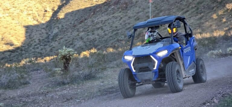 Blue RZR with helmeted riders on a dusty Nevada trail near Las Vegas, cacti, hills—perfect for ATV tours and Ghost Town sightseeing.