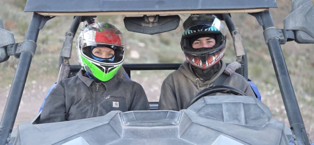 Two people in helmets enjoy a Nevada ATV tour near Las Vegas, riding a RZR with rugged terrain and the Colorado River behind them.