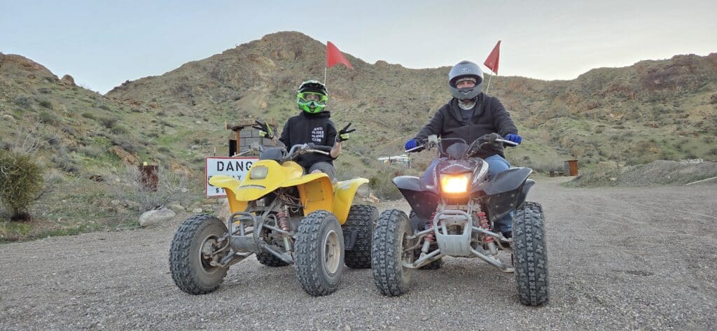 Riders in helmets on yellow-black ATVs tour a rocky Nevada trail near Las Vegas, with red flags and a DANGER sign in view.