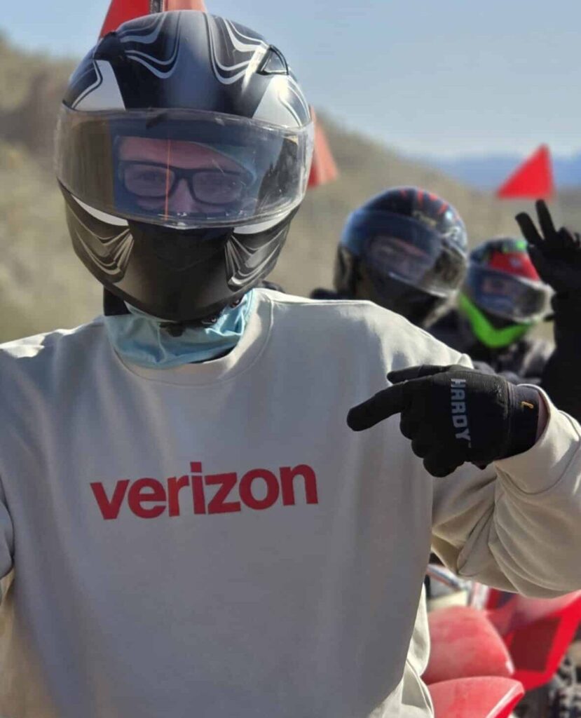 Rider in Verizon gear points at logo on ATV during Nevada ATV tour; group follows with red flags near Las Vegas under sunny skies.
