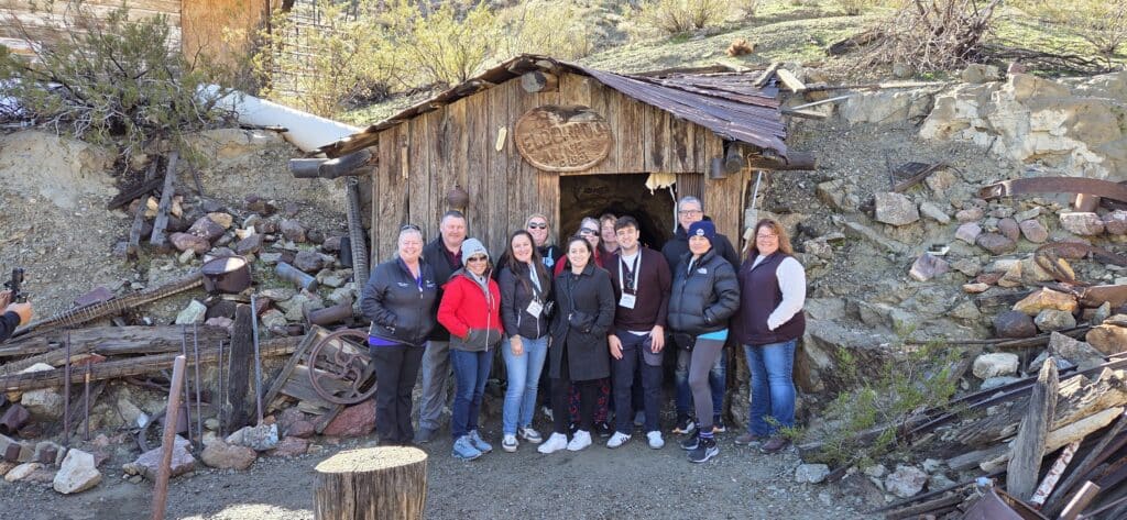 Thirteen people smile outside a rustic Nevada cabin near desert mining gear—perfect for Ghost Town sightseeing or ATV tours.