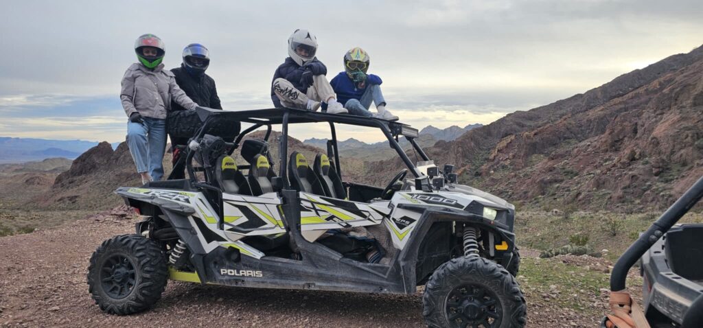 Adventurers in helmets pose by a Polaris ATV on a Nevada trail, enjoying a Las Vegas RZR off-road tour near the Colorado River.