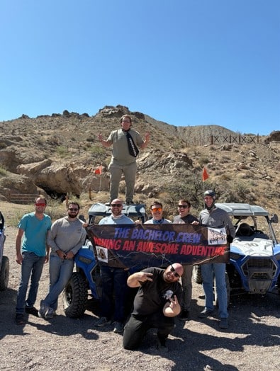 Eight men on a Las Vegas RZR ATV tour pose by rocky hills, banner in hand—ultimate Nevada adventure with Ghost Town sightseeing.