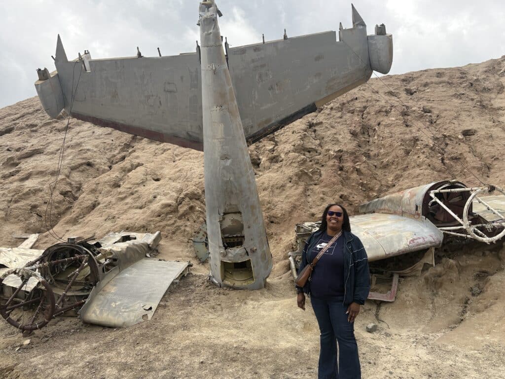 Woman in sunglasses smiles by a weathered airplane wreck on a Nevada hillside, perfect for Ghost Town sightseeing near Las Vegas.