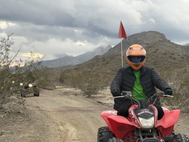 Rider in orange helmet on red ATV explores Nevada desert near Las Vegas with mountains, cloudy skies; ATV tours along Colorado River.