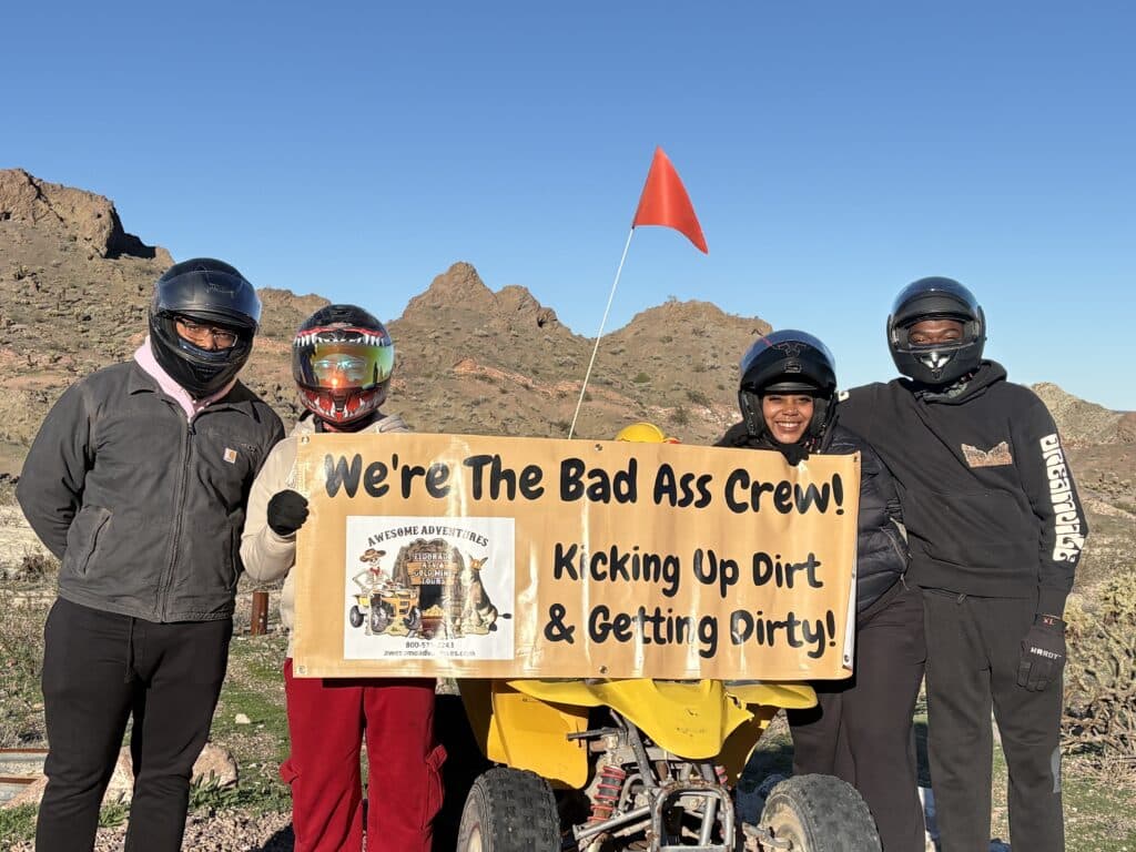 Group in helmets holds "Bad Ass Crew" sign by yellow ATV near Nevada mountains—ATV tour adventure close to Las Vegas.