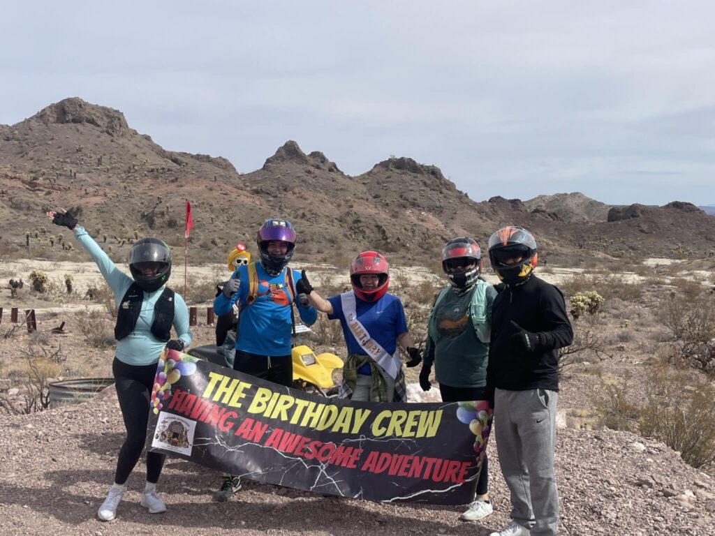 Group in helmets celebrates with a “Birthday Crew” sign before a Nevada mountain—ready for a Las Vegas Ghost Town ATV tour.