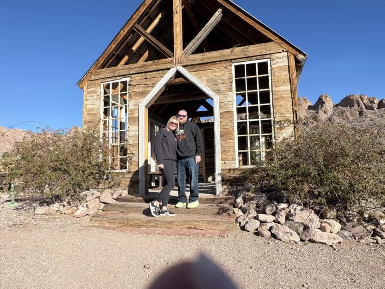 Two people and a small dog smile outside a rustic Nevada ghost town building, perfect for sightseeing near the Colorado River.