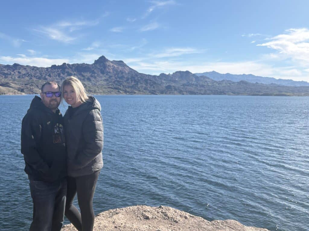 Couple in jackets at Nevada rocky shoreline, blue Colorado River lake, and mountains—Las Vegas Ghost Town sightseeing backdrop.