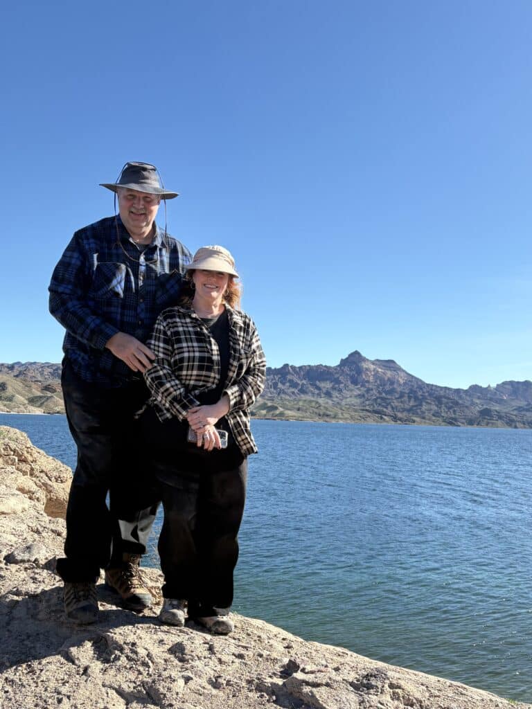 Older couple in hats and plaid by Colorado River near Las Vegas, Nevada, with mountains; perfect for Ghost Town sightseeing tours.