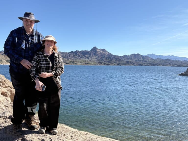 Smiling couple in hats and plaid shirts on Nevada’s rocky shore by the Colorado River near Las Vegas, clear sky, mountain views.
