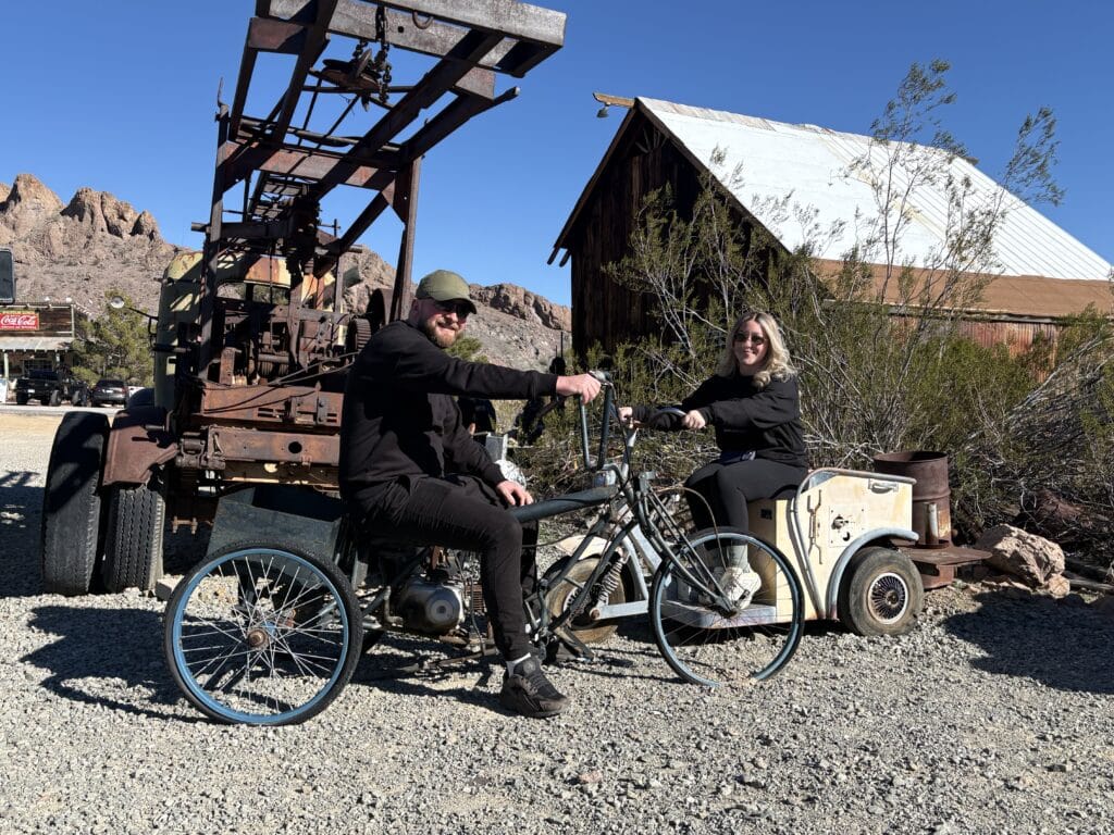 Two people in black on a vintage motorbike in Nevada desert near old shed—perfect for Las Vegas ghost town sightseeing adventures.