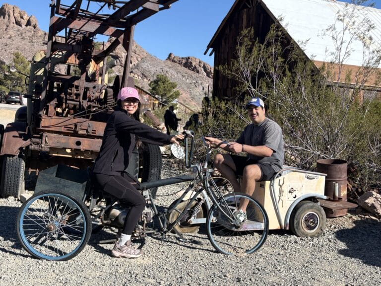 Smiling duo on vintage tricycle in a Nevada ghost town, rustic hills and old barn nearby—perfect for Las Vegas ATV tours.
