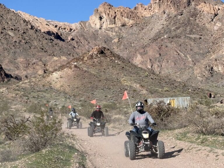 Adventurers ride ATVs with orange flags on a dusty Nevada desert trail near Las Vegas, mountains and clear sky behind.