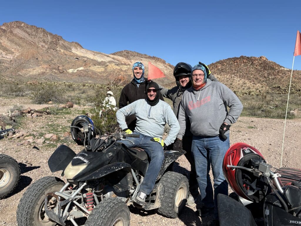 Group of men on an ATV tour near Las Vegas, Nevada, posing on rocky terrain with mountains and clear blue skies in the background.