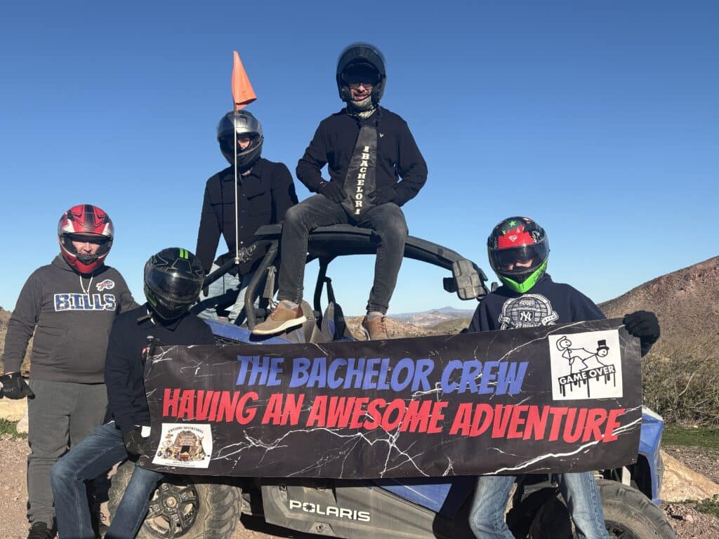 Five helmeted friends pose by an ATV in the Nevada desert near Las Vegas, banner reads THE BACHELOR CREW HAVING AN AWESOME ADVENTURE.