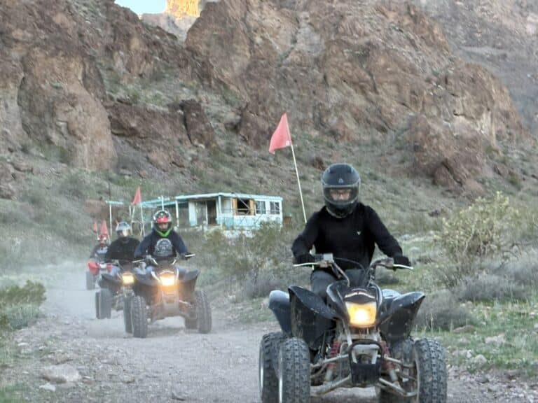 ATV riders explore a dusty Nevada trail near Las Vegas with red flags, passing a ghost town building on an exciting off-road tour.
