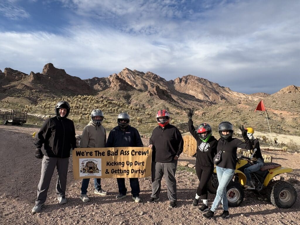 Group in helmets holding “We’re The Bad Ass Crew!” sign on a Nevada ATV tour near Las Vegas, with mountains in the background.